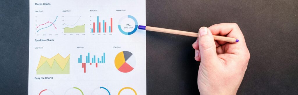 Hand holding pencil reviewing colorful data charts on desk with laptop.